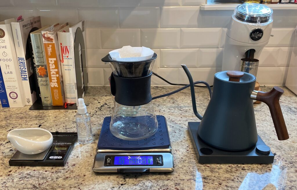 Coffee dosing tray on a small scale beside a V60 dripper on a second digital scale, kettle and grinder.